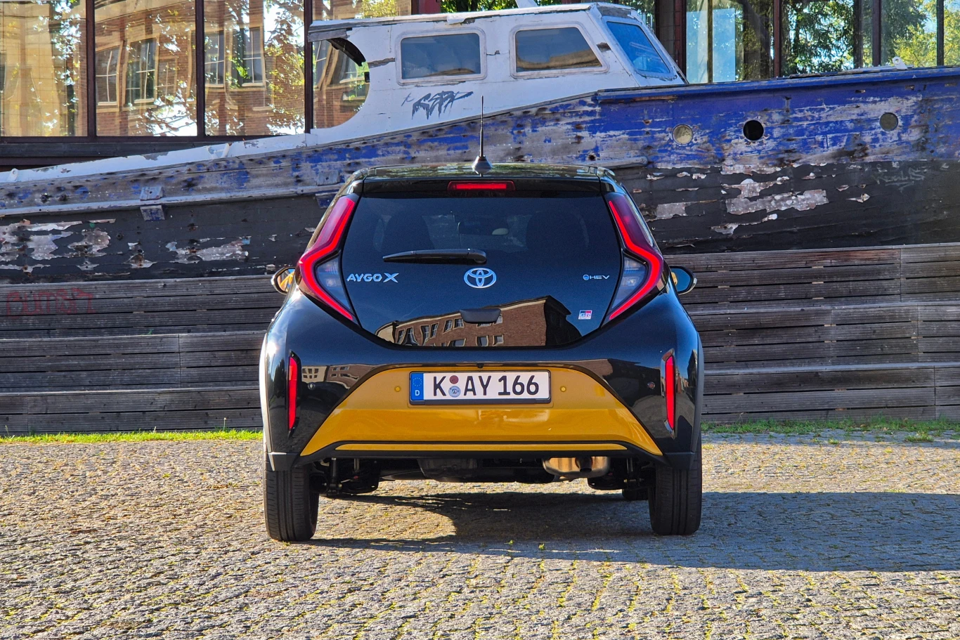 Czarny i żółty samochód osobowy marki Toyota Aygo X sfotografowany z tyłu, zaparkowany na żwirowej powierzchni, w tle widoczny stary, zniszczony drewniany kuter rybacki.