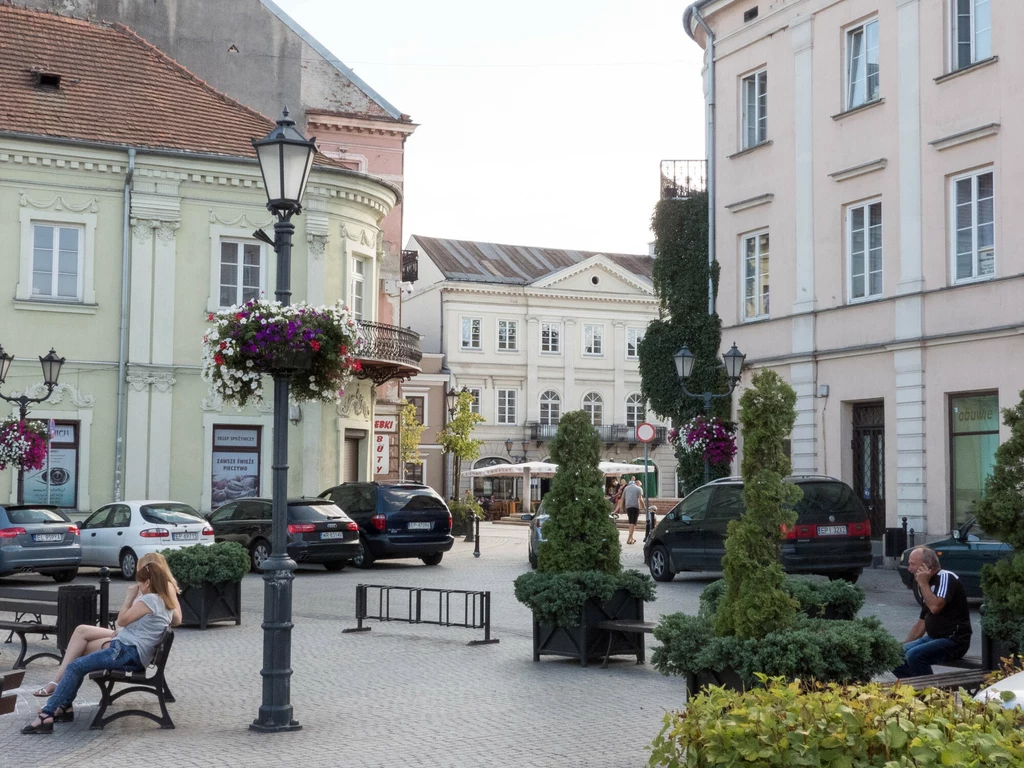 Urokliwy skwer miejski otoczony zabytkowymi kamienicami, ozdobionymi kwiatami w wiszących donicach oraz krzewami w donicach. Na ławkach siedzą spokojnie dwie osoby, a w tle zaparkowane są samochody.