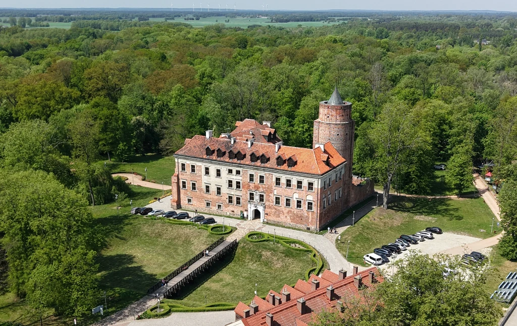 Zamek o czerwonych dachach i wieży obronnej otoczony zielonym lasem, z widocznym dziedzińcem oraz zaparkowanymi samochodami po jednej stronie. W tle rozciąga się gęsty las i pola uprawne, a przed zamkiem zadbane alejki i fragment nowoczesnych zabudowań.