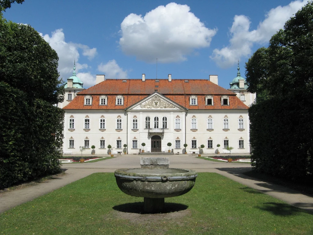 Barokowy pałac z czerwonym dachem, symetryczną fasadą i licznymi oknami, przed którym znajduje się kamienna fontanna otoczona zielonym trawnikiem i przyciętymi żywopłotami, całość w słoneczny dzień z kilkoma chmurami na niebie
