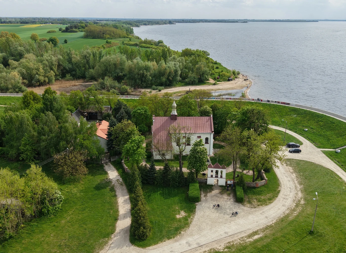 Kościół o białych ścianach i czerwonym dachu otoczony zielenią, położony niedaleko wybrzeża jeziora, wokół widoczne alejki oraz drzewa, z widokiem na spokojną wodę i pola w tle.