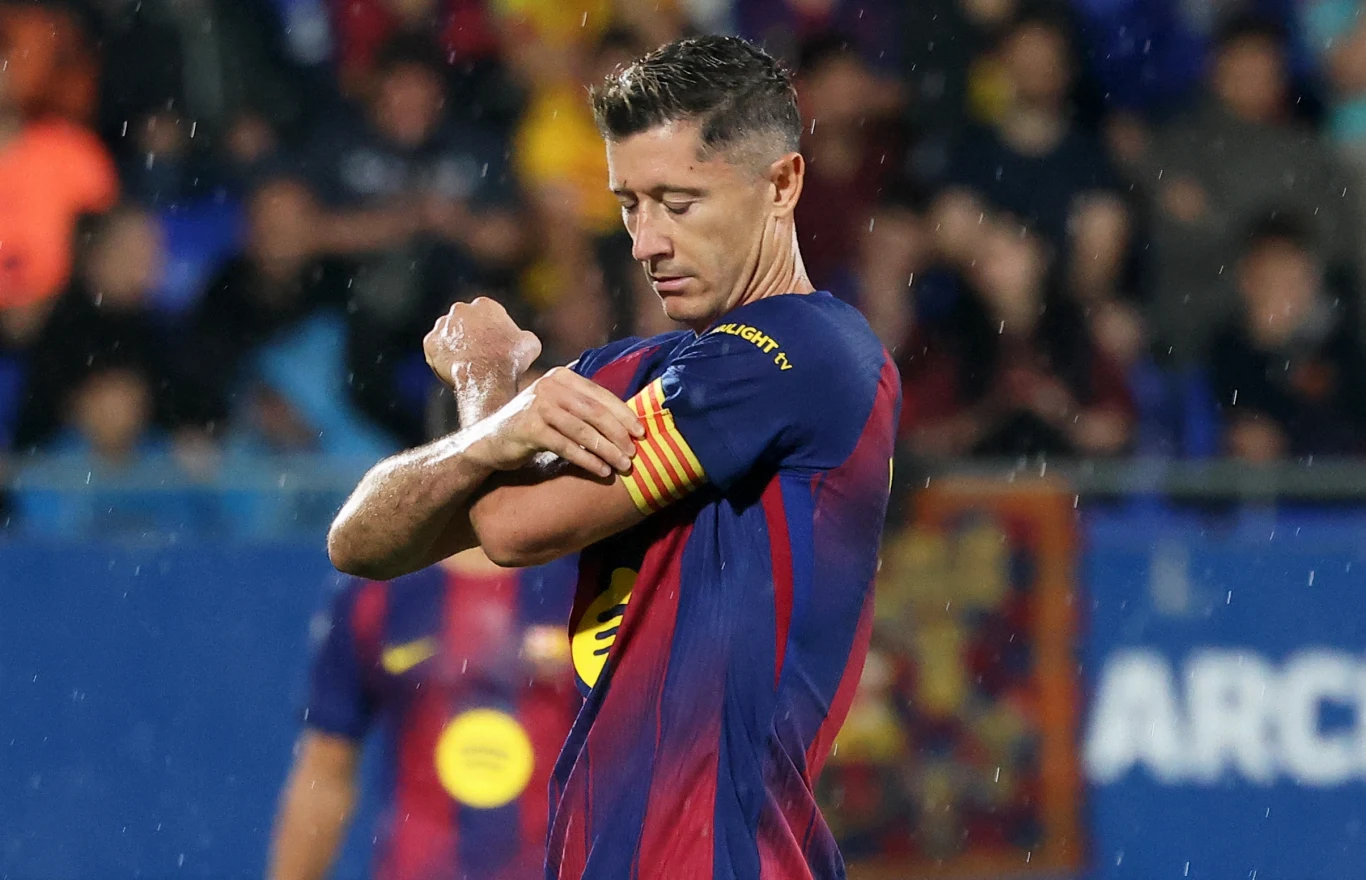 Robert Lewandowski Piłkarz w stroju klubu FC Barcelona zakłada opaskę kapitana na ramieniu podczas meczu, w tle rozmazana publiczność stadionowa oraz padający deszcz
