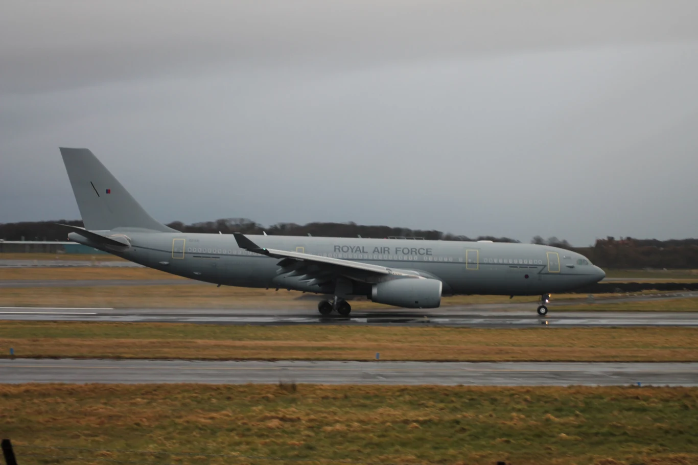 Tankowiec Airbus Voyager KC Mk2 Voyager w bazie lotniczej RAF Prestwick Samolot wojskowy Royal Air Force w barwach szarości kołuje po pasie startowym na lotnisku, w tle widoczne są drzewa i niskie wzgórza przy pochmurnym niebie.