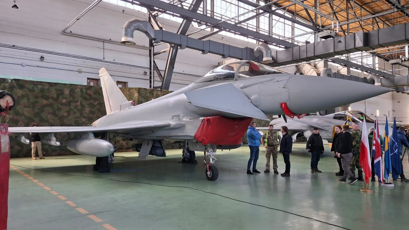 Brytyjski Eurofighter Typohoon FGR4 w 22. Bazie Lotnictwa Taktycznego w Malborku Myśliwiec wojskowy ustawiony w hangarze, wokół samolotu stoi grupa ludzi, w tym wojskowi i cywile, w tle widoczne flagi różnych państw, a nad głową żelazna konstrukcja dachu.