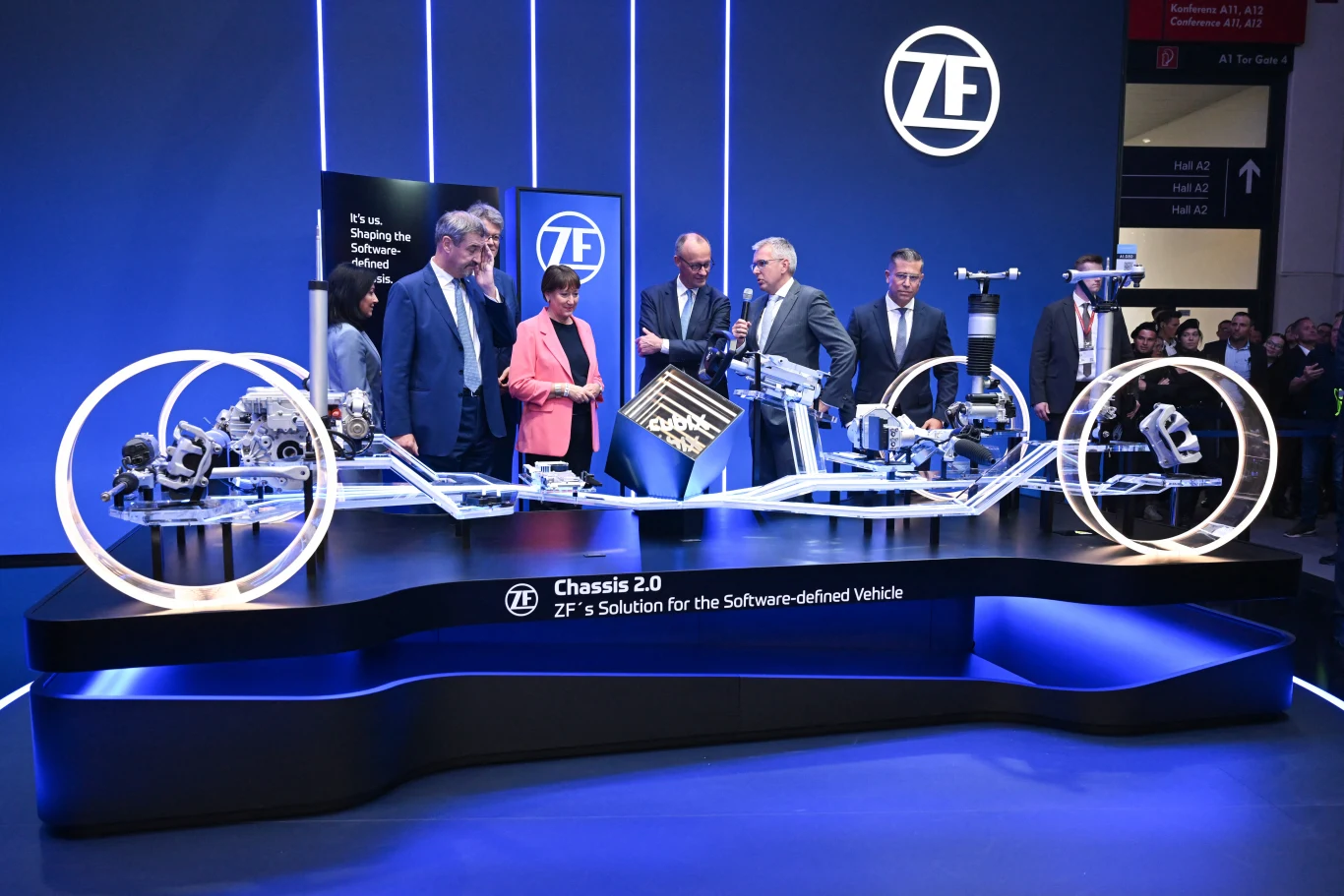 Dyrektor generalny ZF Holger Klein w kanclerzem Niemiec Friedrichem Merzema podczas IAA Mobility w Monachium Grupa osób w eleganckich ubraniach omawia nowoczesny model podwozia samochodu wyeksponowany na podświetlonym stanowisku wystawowym, w tle logo firmy ZF i prezentacja multimedialna.