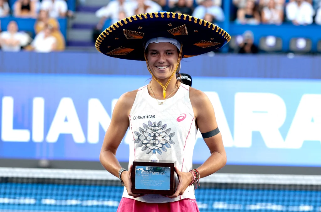 Uśmiechnięta tenisistka w sportowym stroju i ozdobnym sombrero trzyma w dłoniach nagrodę na tle kortu tenisowego oraz widowni.