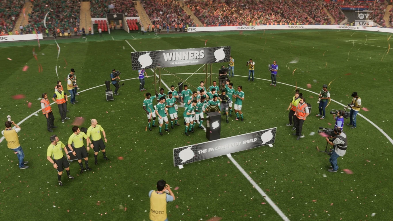 EA Sports FC 26 Zwycięska drużyna piłkarska w zielonych strojach świętuje na stadionie pod napisem WINNERS, otoczona przez fotoreporterów, kamerzystów oraz sędziów, wśród konfetti i wiwatującej publiczności na trybunach.