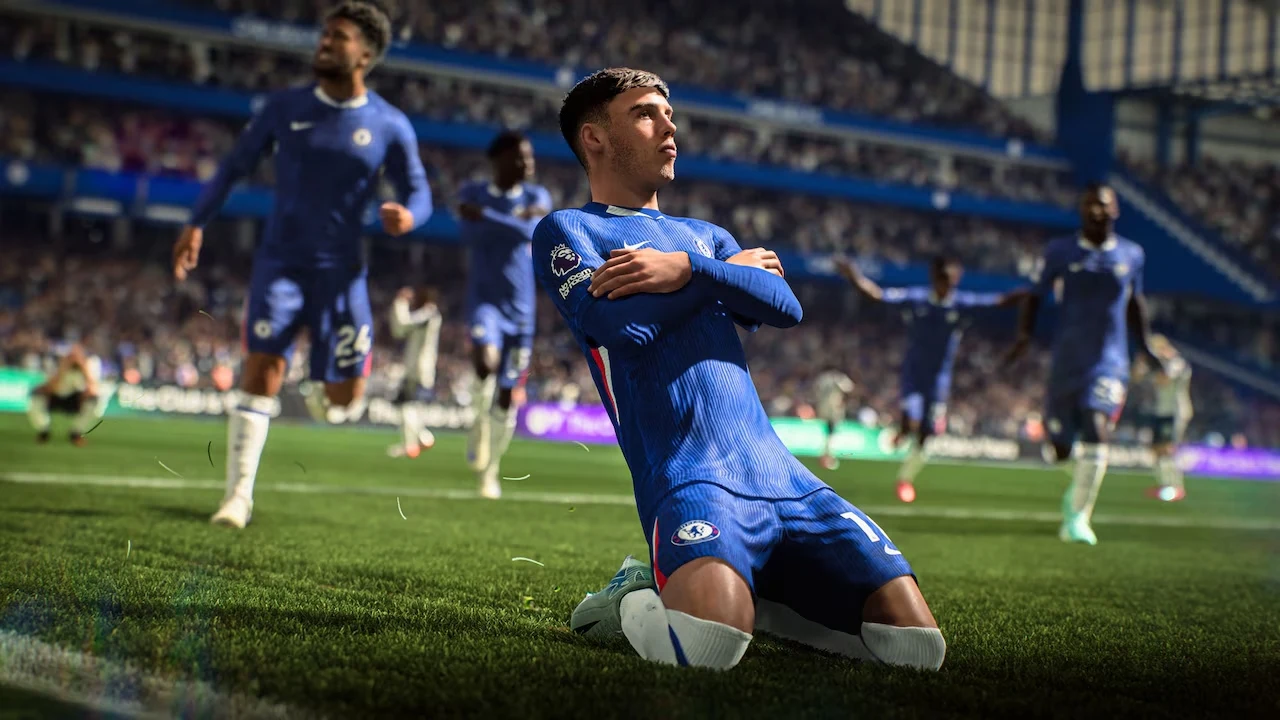 EA Sports FC 26 Piłkarz w niebieskim stroju świętuje zdobycie gola na trawie stadionu, klęcząc i z szeroko rozłożonymi ramionami, w tle widać kibiców oraz innych zawodników biegnących w jego stronę.