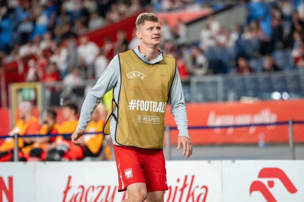 Paweł Dawidowicz Piłkarz ubrany w sportową kamizelkę treningową i spodenki z godłem Polski przechadza się po murawie stadionu, w tle kibice na trybunach i fragment ławki rezerwowych.