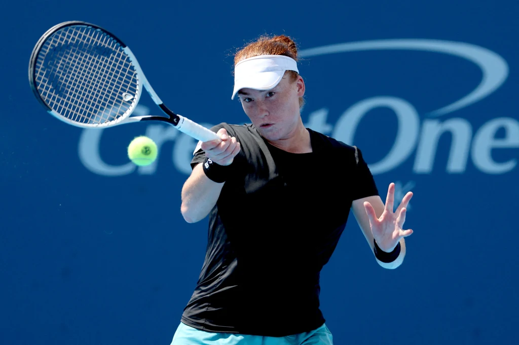 Tenisistka w czarnej koszulce i białej daszce odbija piłkę tenisową podczas meczu na tle niebieskiej ściany z logo reklamowym.