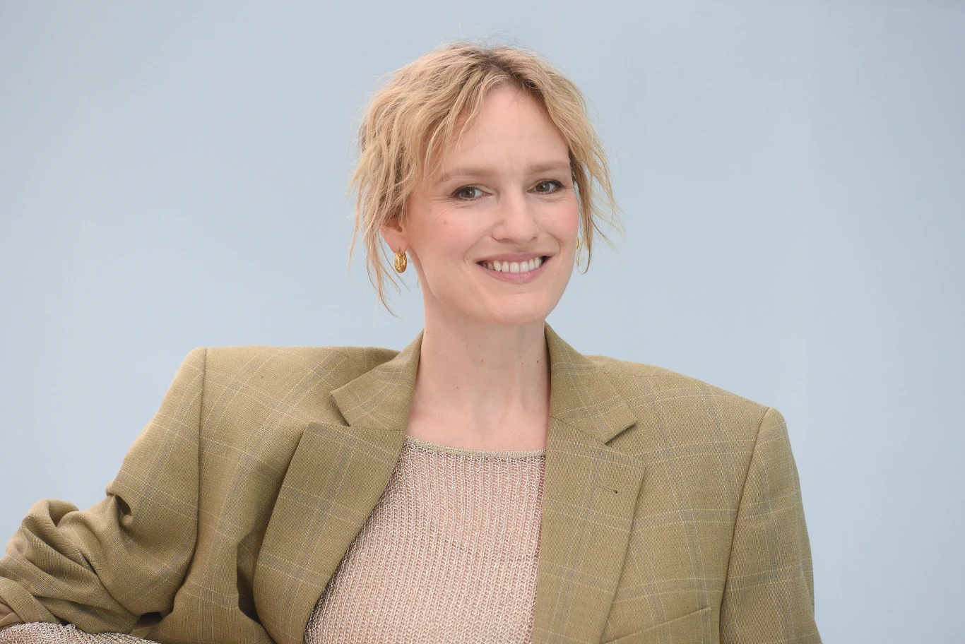 Bovska Kobieta o blond włosach w krótkiej fryzurze uśmiecha się, ubrana w beżowy sweter i marynarkę w stonowanym kolorze na jasnym tle.