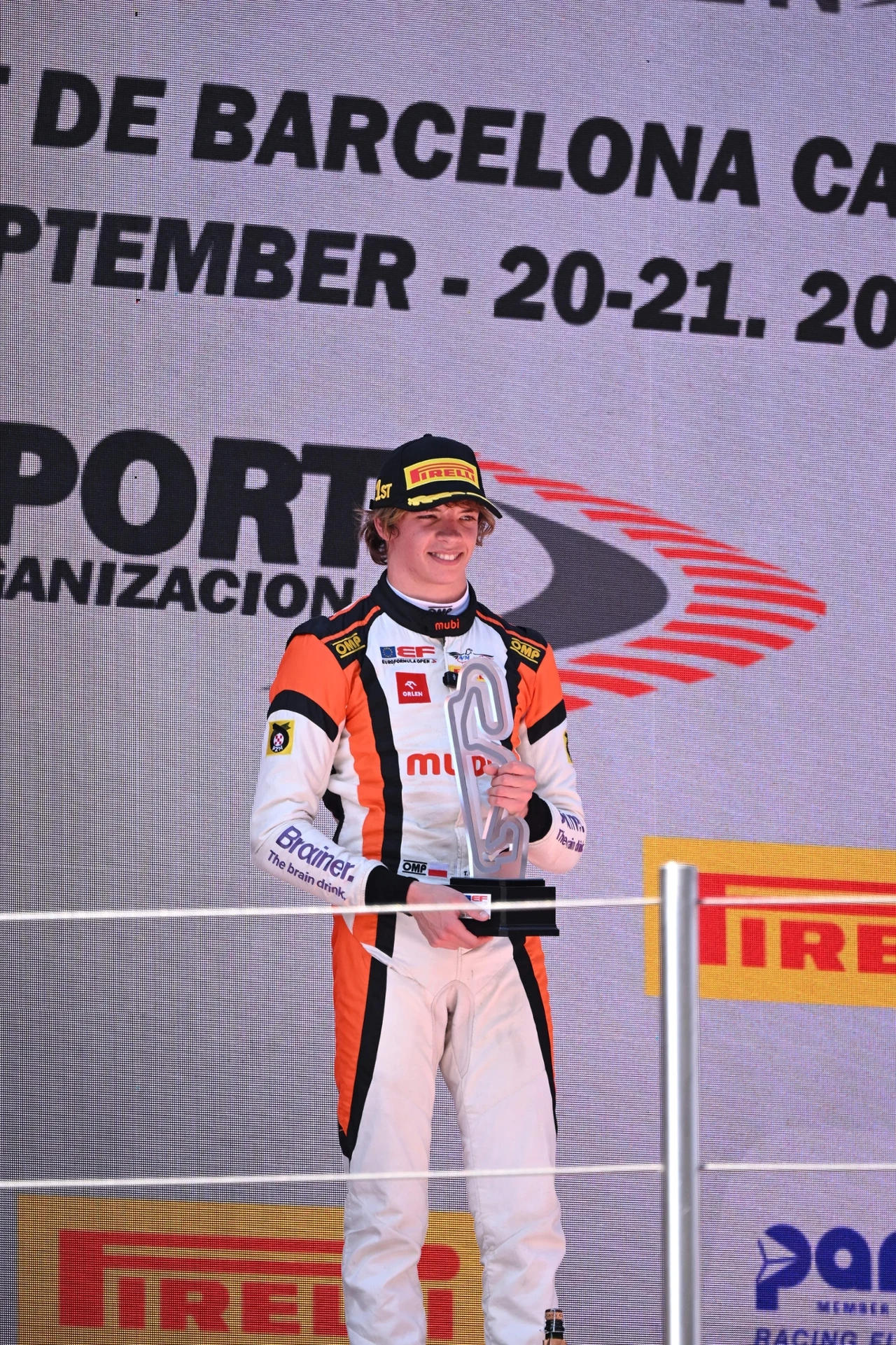 Fot. archiwum: Tymek Kucharczyk Kierowca wyścigowy w stroju sportowym stojący na podium trzyma w rękach puchar, w tle widoczne są logotypy sponsorów oraz nazwa toru i data wydarzenia.