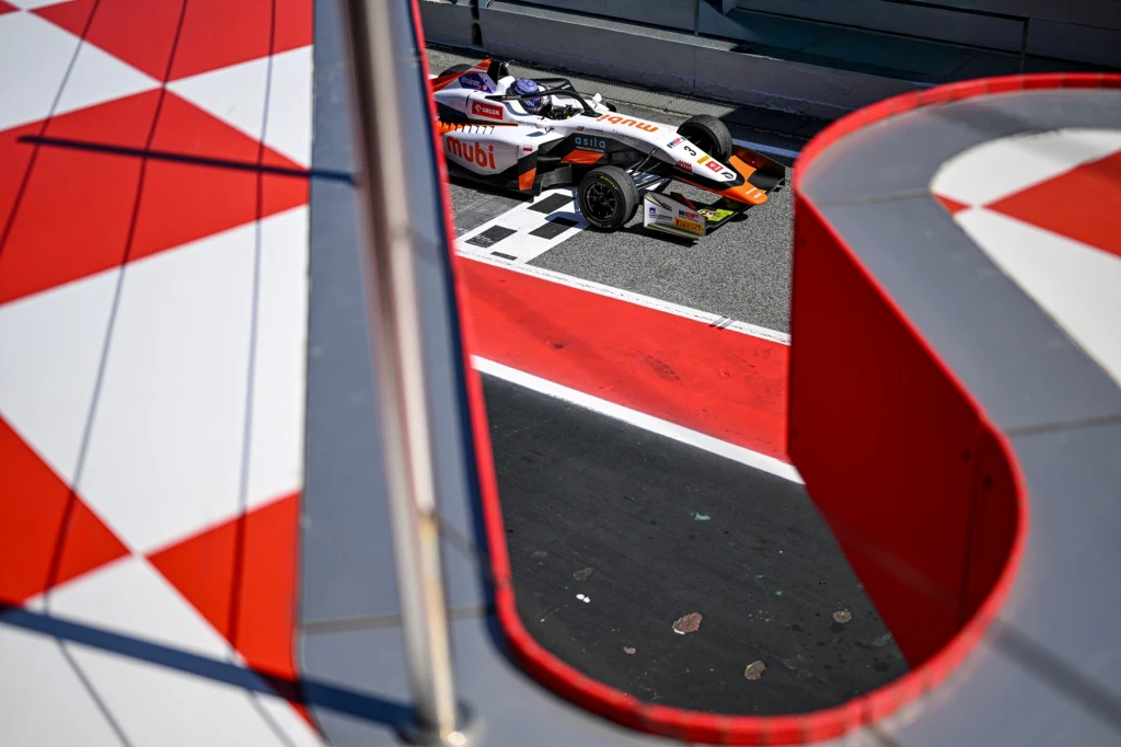 Fot. archiwum: Tymek Kucharczyk Bolid wyścigowy przejeżdżający przez linię na torze, widoczny z ciekawej perspektywy przez element konstrukcyjny z biało-czerwonym wzorem.