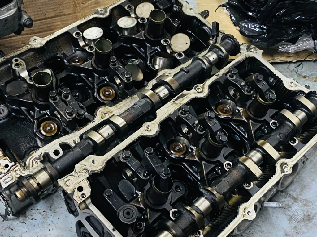 Osady i nagar w silniku – efekt braku regularnego płukania silnika Dwie głowice silnika samochodowego z widocznymi wałkami rozrządu oraz elementami zaworów, częściowo rozebrane i umieszczone na stole warsztatowym.