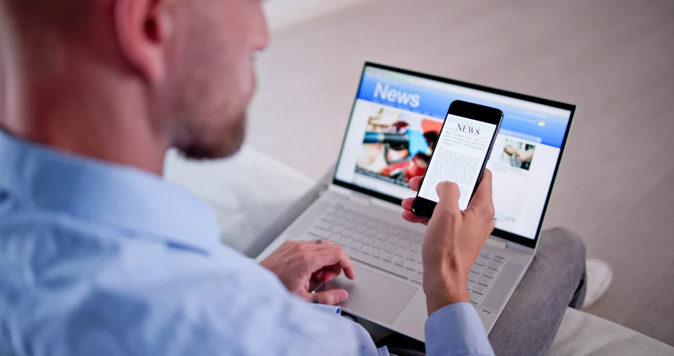 Większość ruchu w Internecie tworzą obecnie nie ludzie, lecz boty. Mężczyzna siedzi z laptopem na kolanach, sprawdzając wiadomości na smartfonie, na ekranie komputera widoczna sekcja z informacjami.