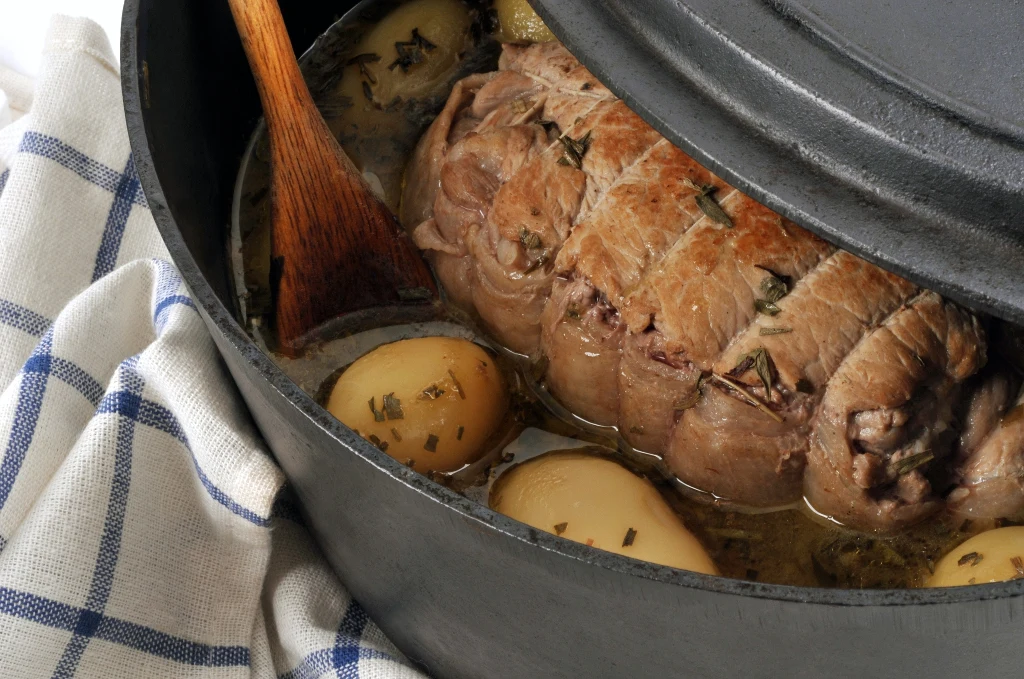 Duszona pieczeń wołowa w żeliwnym garnku z ziemniakami i przyprawami, na stole przykrytym kuchenną ściereczką w niebieską kratę, drewniana łyżka dołączona do potrawy.
