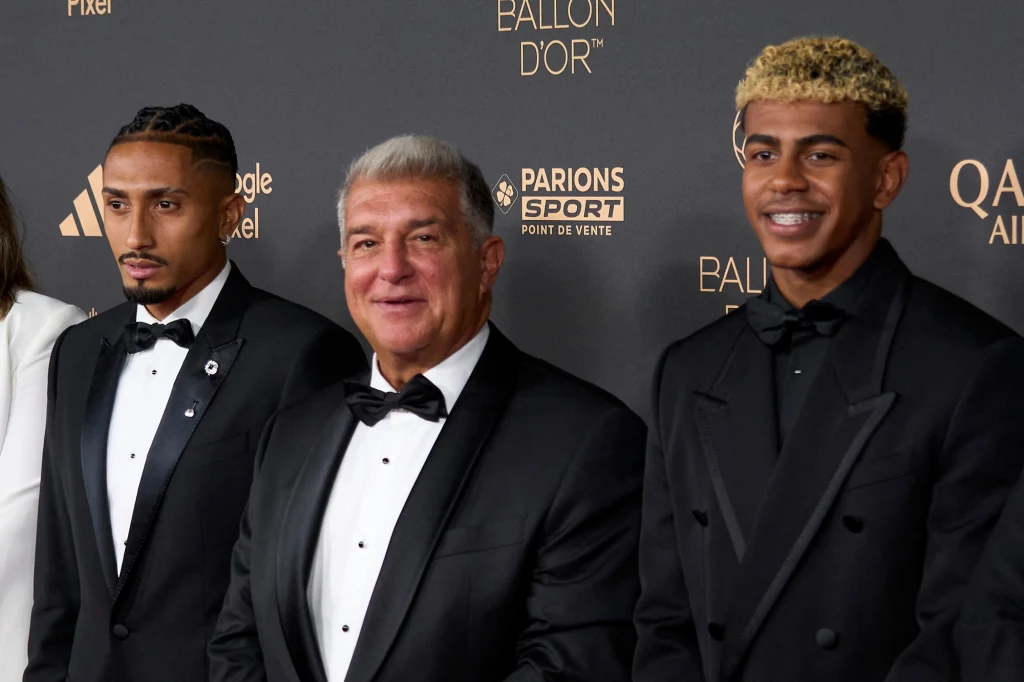 Raphinha, Joan Laporta i Lamine Yamal na gali Złotej Piłki Trzech elegancko ubranych mężczyzn w smokingu pozuje wspólnie na tle ścianki z logotypami partnerów oraz napisem Ballon d'Or, tło wskazuje na galę lub oficjalne wydarzenie sportowe.