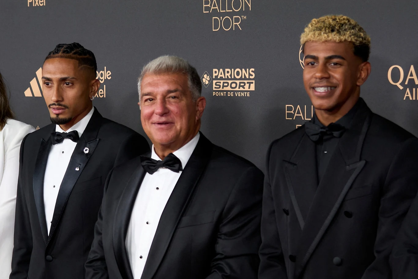 Trzech elegancko ubranych mężczyzn w smokingu pozuje wspólnie na tle ścianki z logotypami partnerów oraz napisem Ballon d'Or, tło wskazuje na galę lub oficjalne wydarzenie sportowe.