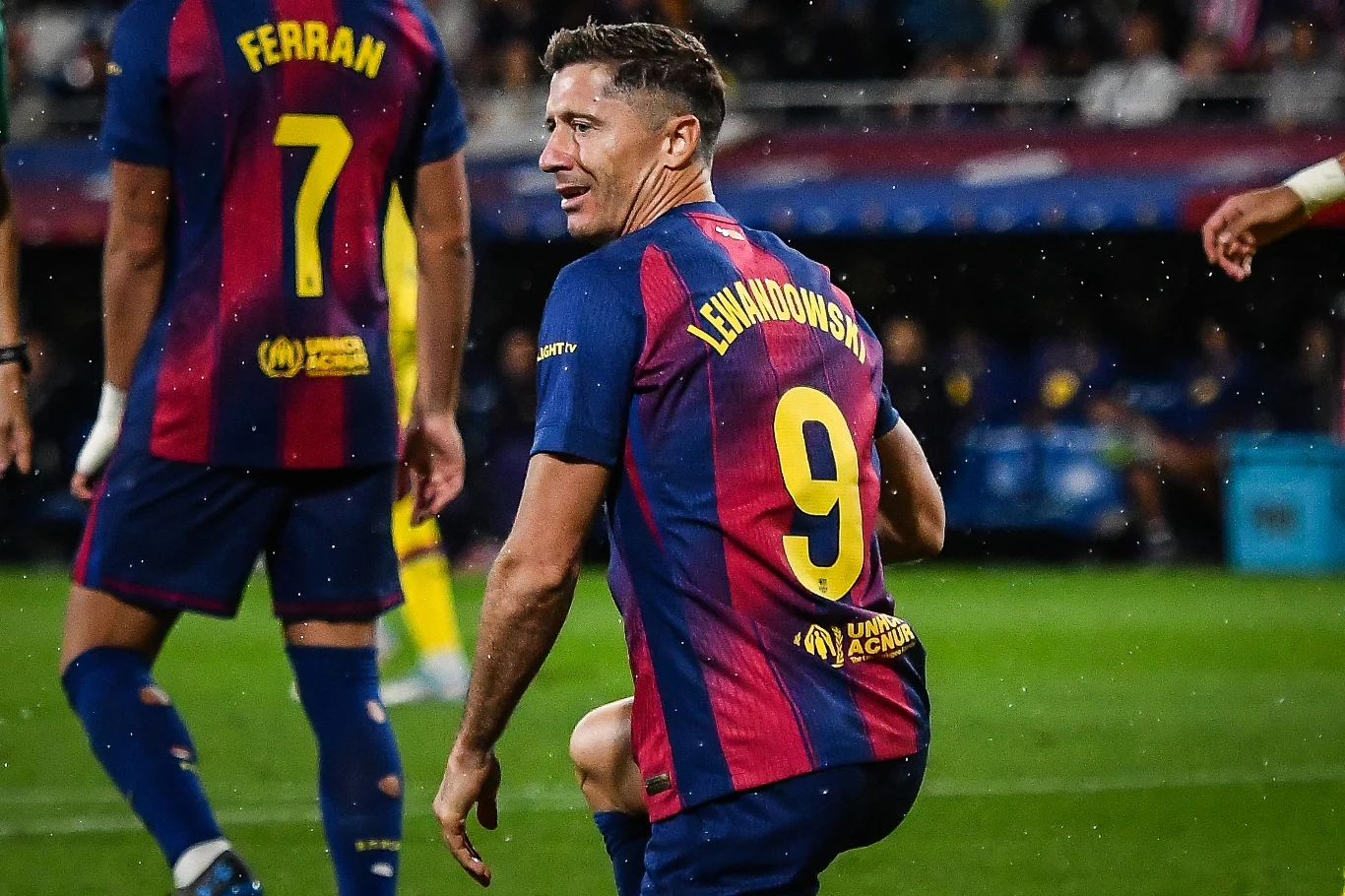 Piłkarz drużyny FC Barcelona w koszulce z numerem 9 z nazwiskiem Lewandowski klęczy na murawie podczas meczu, obok znajduje się inny zawodnik w stroju tego samego zespołu z numerem 7