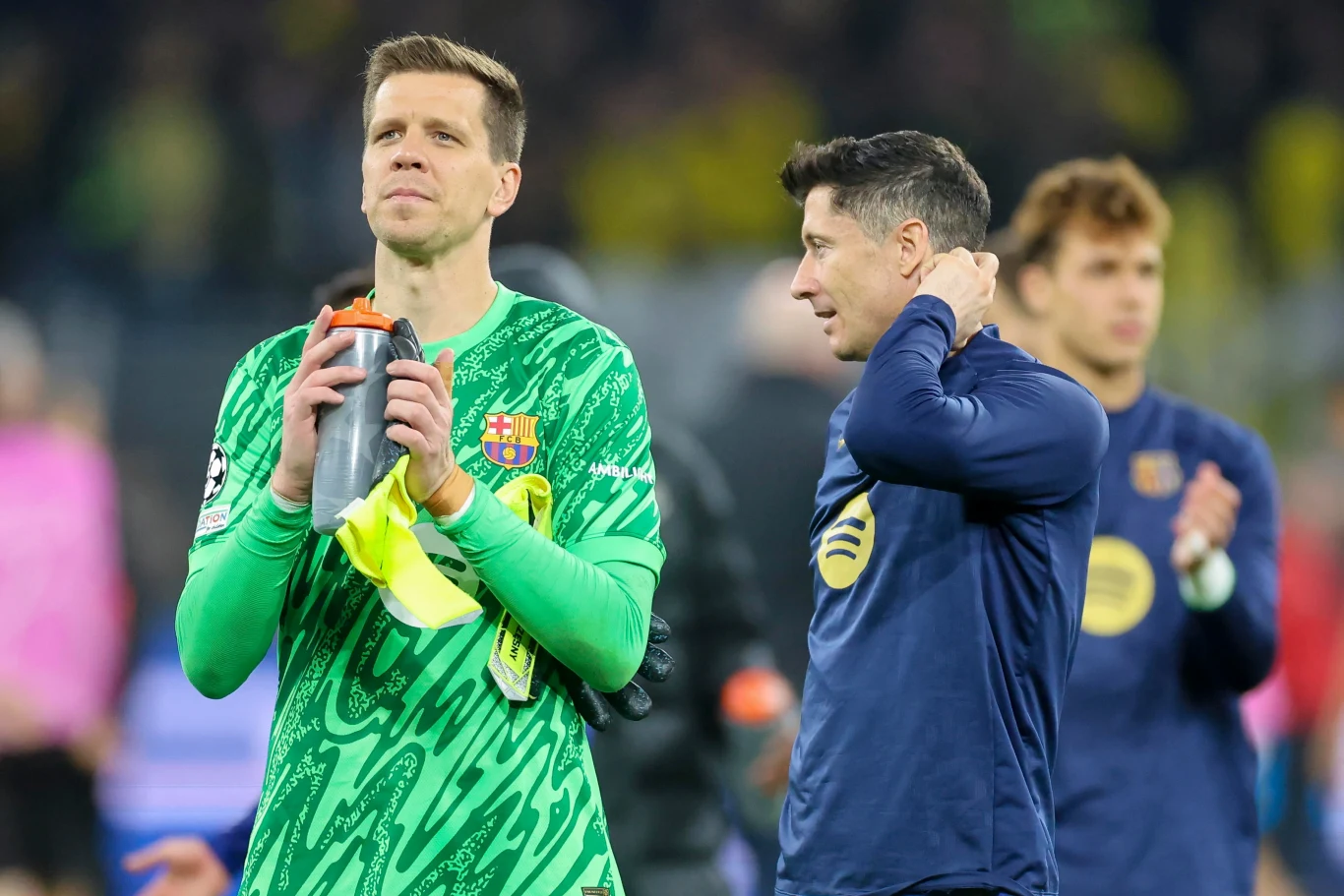 Dwóch piłkarzy na murawie stadionu po zakończonym meczu, jeden z nich w zielonym stroju bramkarskim trzyma butelkę i żółty ręcznik, drugi w granatowej bluzie poprawia kołnierzyk, w tle pozostali zawodnicy i rozmyta publiczność.