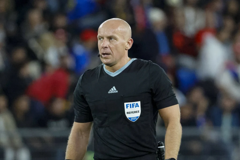 Szymon Marciniak Łysy mężczyzna w czarnym stroju sędziowskim z emblematem FIFA, stojący na tle rozmytej widowni podczas wydarzenia sportowego.