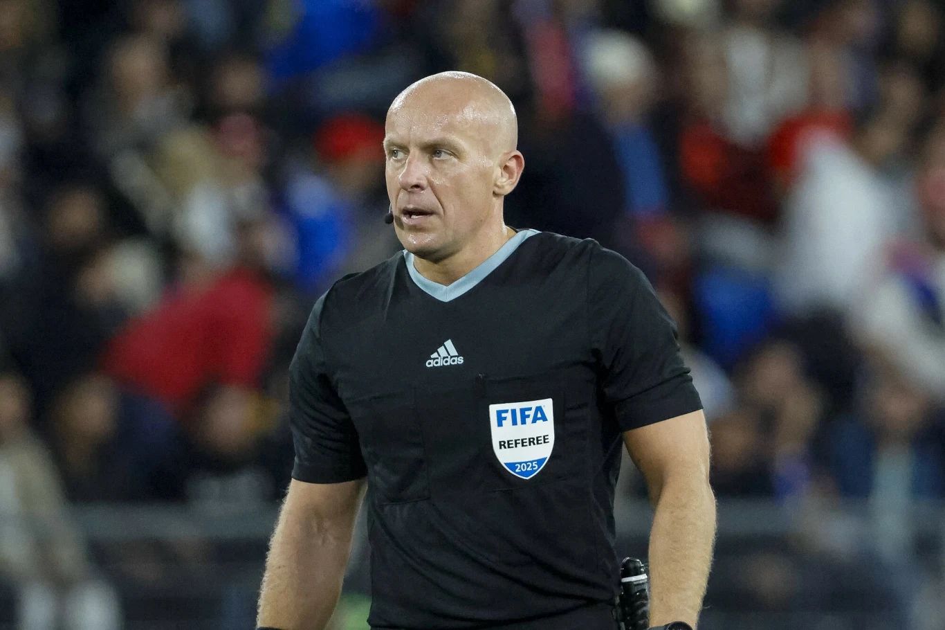 Szymon Marciniak Łysy mężczyzna w czarnym stroju sędziowskim z emblematem FIFA, stojący na tle rozmytej widowni podczas wydarzenia sportowego.