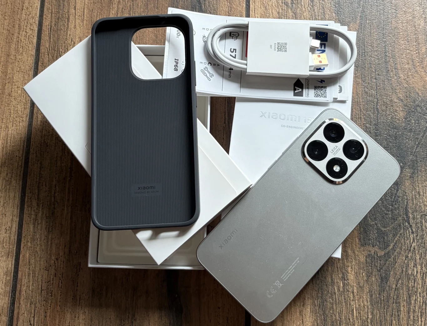 Nowoczesny smartfon marki Xiaomi leżący obok otwartego opakowania, do którego dołączone są silikonowe etui, kabel USB oraz dokumentacja. Wszystko umieszczone na drewnianej powierzchni.