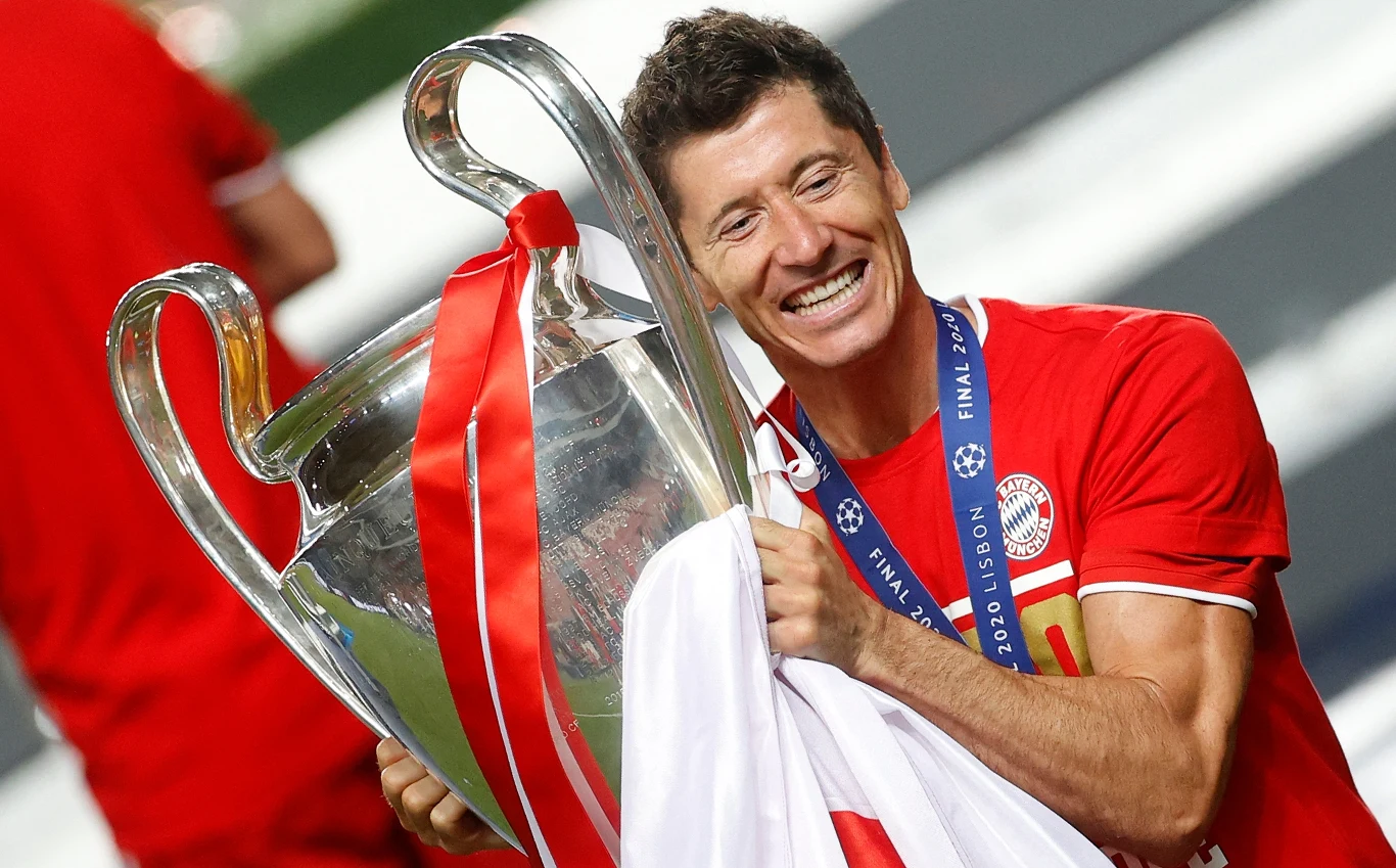 Piłkarz w czerwonej koszulce trzyma duży puchar Ligi Mistrzów, szeroko się uśmiecha, zawieszony medal na szyi, w tle nieostre postacie w sportowych strojach.