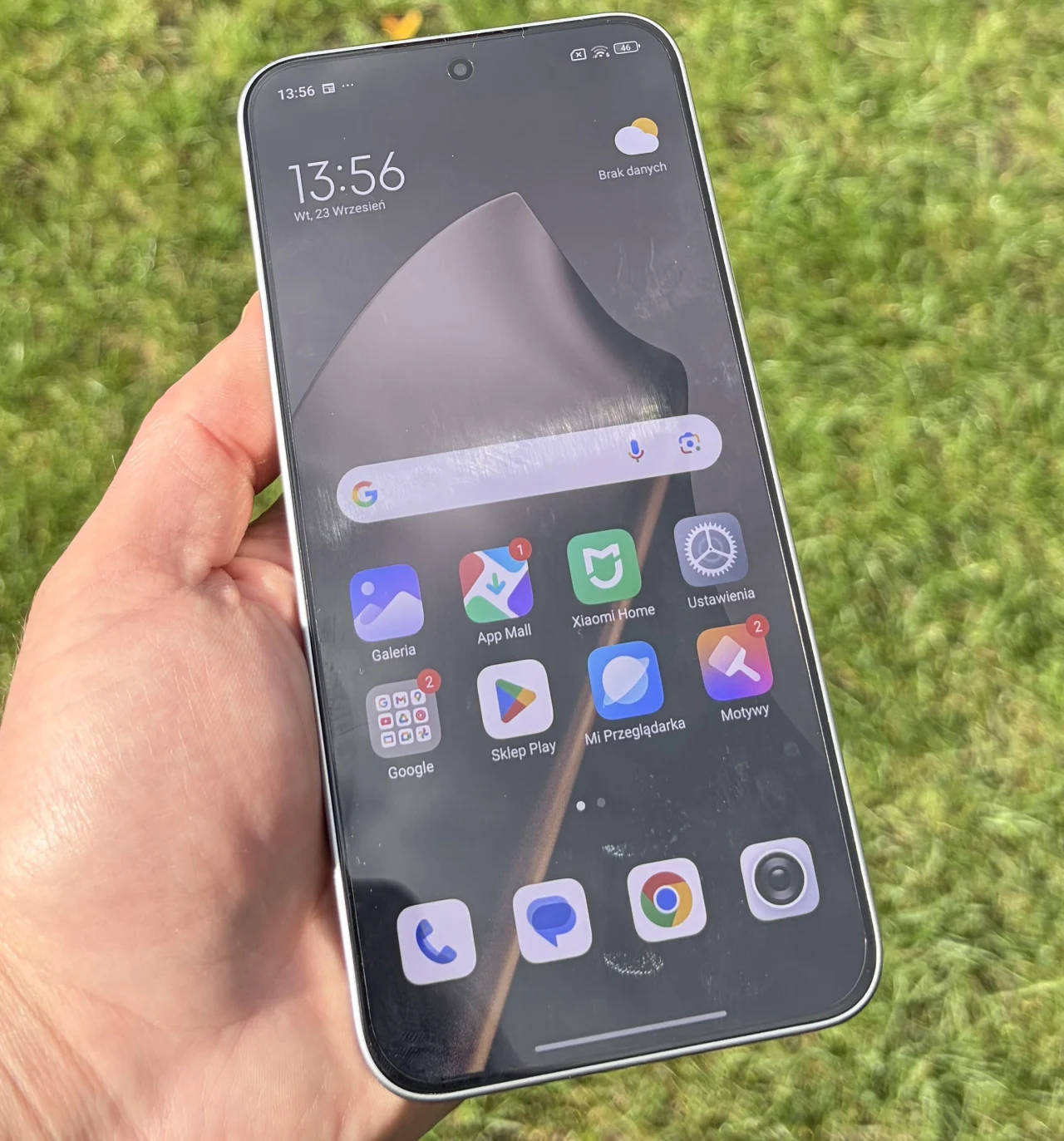 Nowoczesny smartfon trzymany w lewej ręce na tle zielonej trawy, ekran telefonu pokazuje aplikacje oraz powiadomienia, górna część wyświetlacza prezentuje godzinę i datę.