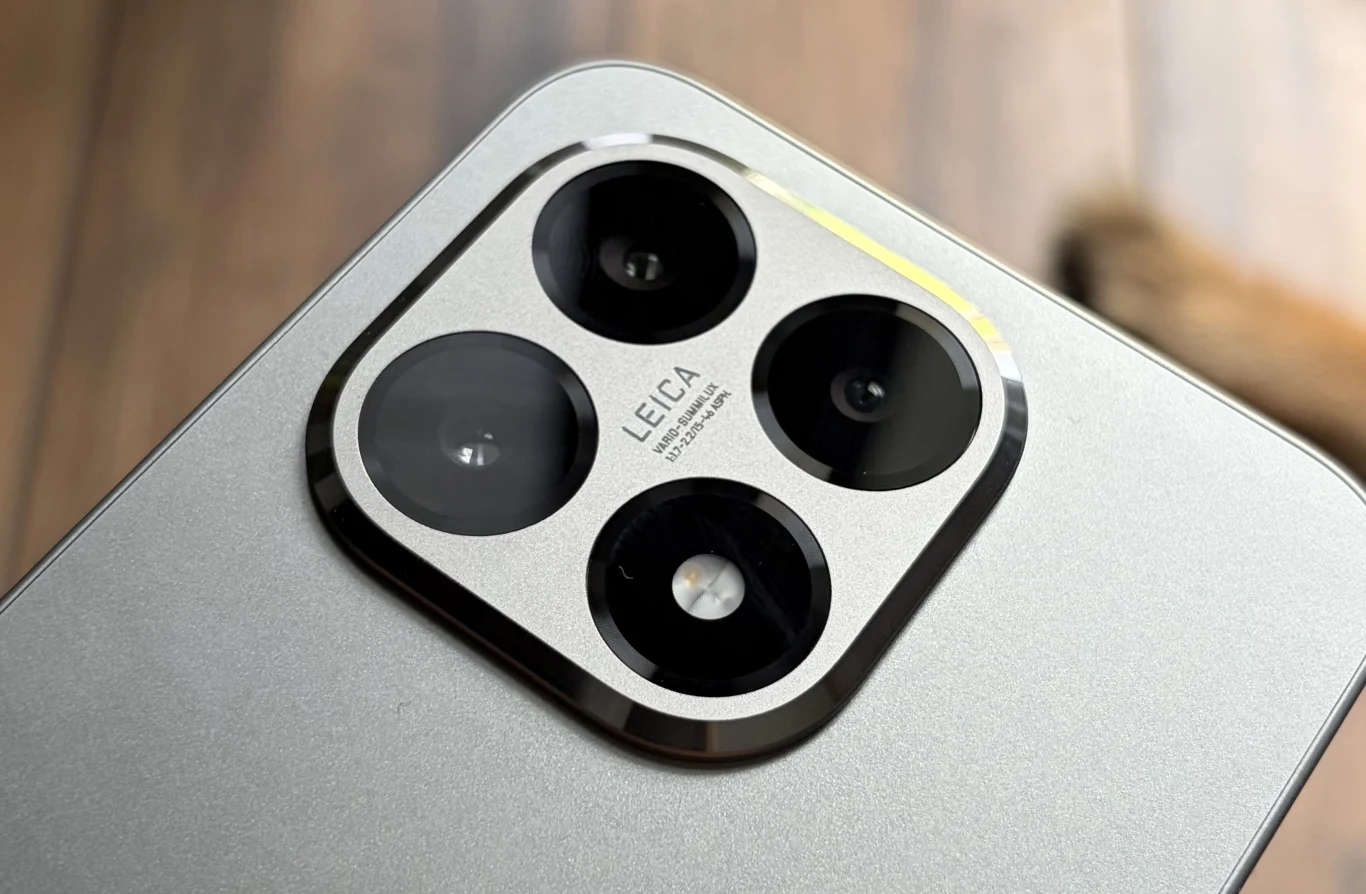 Metaliczny moduł aparatu z trzema dużymi obiektywami oraz logotypem Leica na prostokątnej obudowie smartfona, na tle rozmazanego drewnianego podłoża.