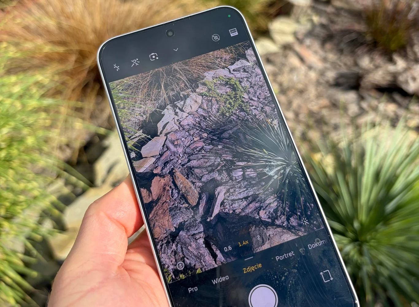 Dłoń trzymająca smartfon, na ekranie którego widać aplikację aparatu fotograficznego skierowaną na skalistą powierzchnię z roślinami, w tle podobny krajobraz z suchymi trawami i kamieniami.