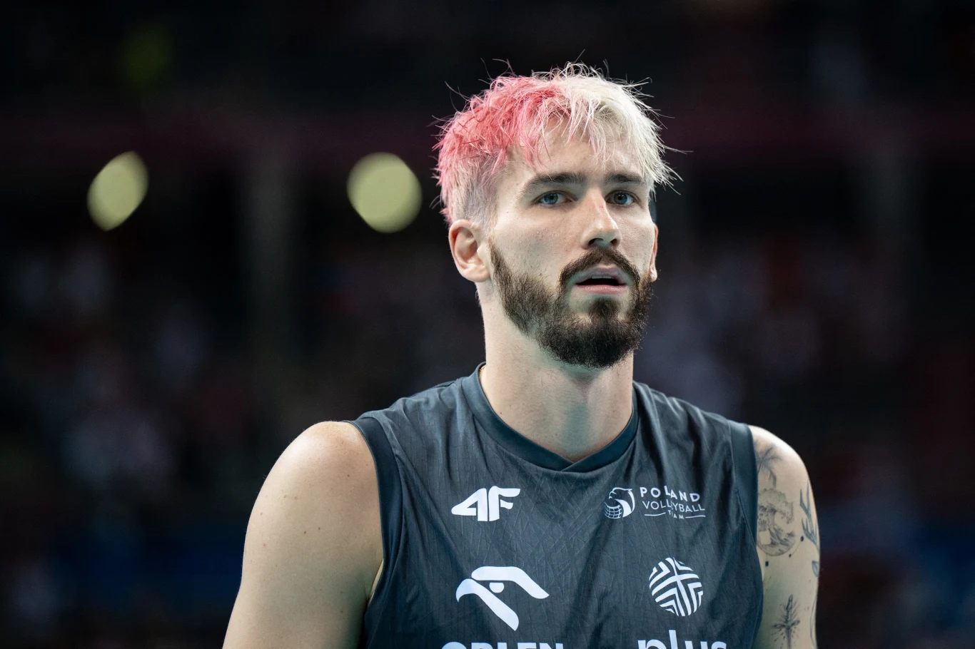 Tomasz Fornal Siatkarz w ciemnym stroju reprezentacyjnym Polski z jasnymi włosami farbowanymi na różowo po jednej stronie, skupiony, stojący na boisku podczas meczu sportowego.