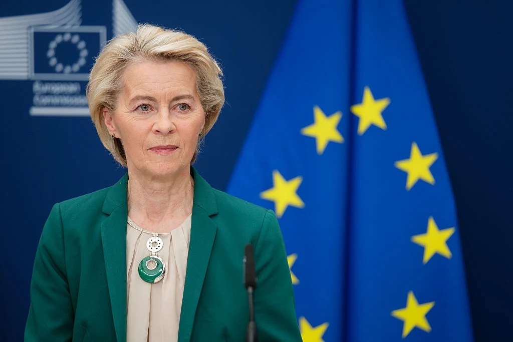 Przewodnicząca KE Ursula von der Leyen Kobieta w zielonym żakiecie stoi przed mikrofonem na tle niebieskiej flagi Unii Europejskiej z żółtymi gwiazdami oraz logo instytucji europejskiej.