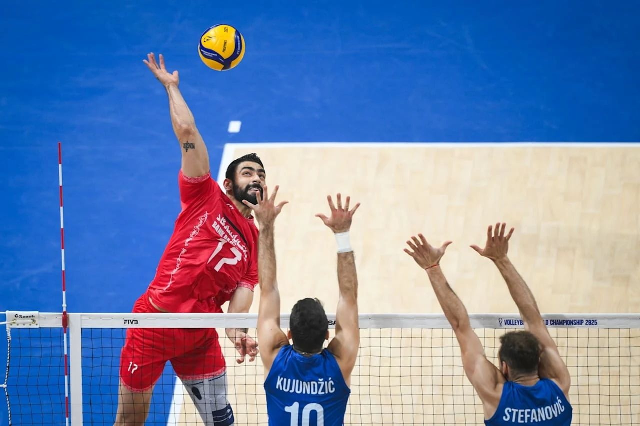 Siatkarz w czerwonym stroju atakuje piłkę nad siatką, podczas gdy dwóch zawodników w niebieskich koszulkach próbuje zablokować jego uderzenie, piłka unosi się nad boiskiem z niebieskim tłem.