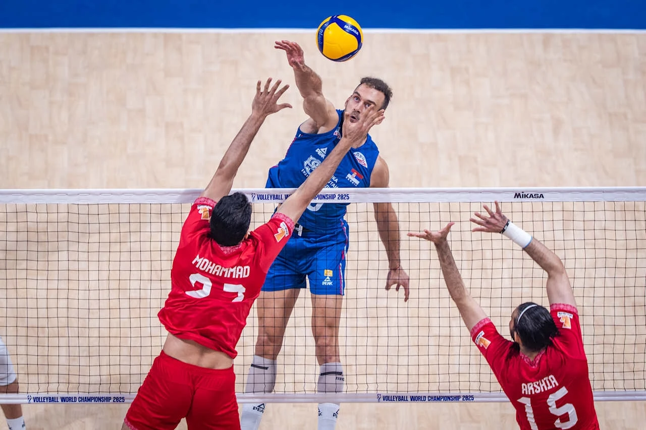 Zawodnik w niebieskim stroju wykonuje atak nad siatką podczas meczu siatkówki, dwaj zawodnicy w czerwonych strojach próbują go zablokować, piłka znajduje się tuż nad siatką