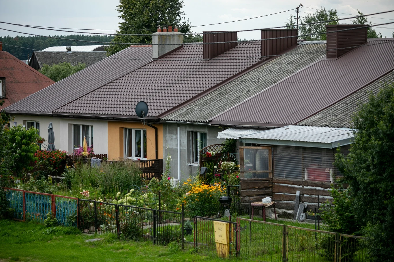 Wymiany dachu z azbestu w gospodarstwach rolnych. Termin ma zostać wydłużony Szereg domów jednorodzinnych z małymi ogrodami, kwiatami i ogrodzeniem w pierwszym planie, dachy budynków mają różne stopnie zużycia, na jednym z domów widoczna jest satelitarna antena.