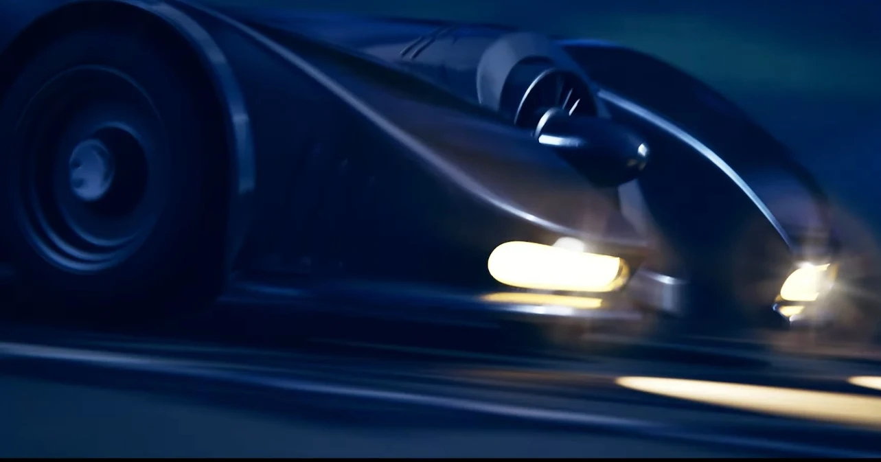 Batmobil wjeżdża do uniwersum Fortnite'a Wyścigowy samochód sportowy poruszający się z dużą prędkością po torze, dynamicznie oświetlony reflektorami, z rozmytym tłem sugerującym szybki ruch.