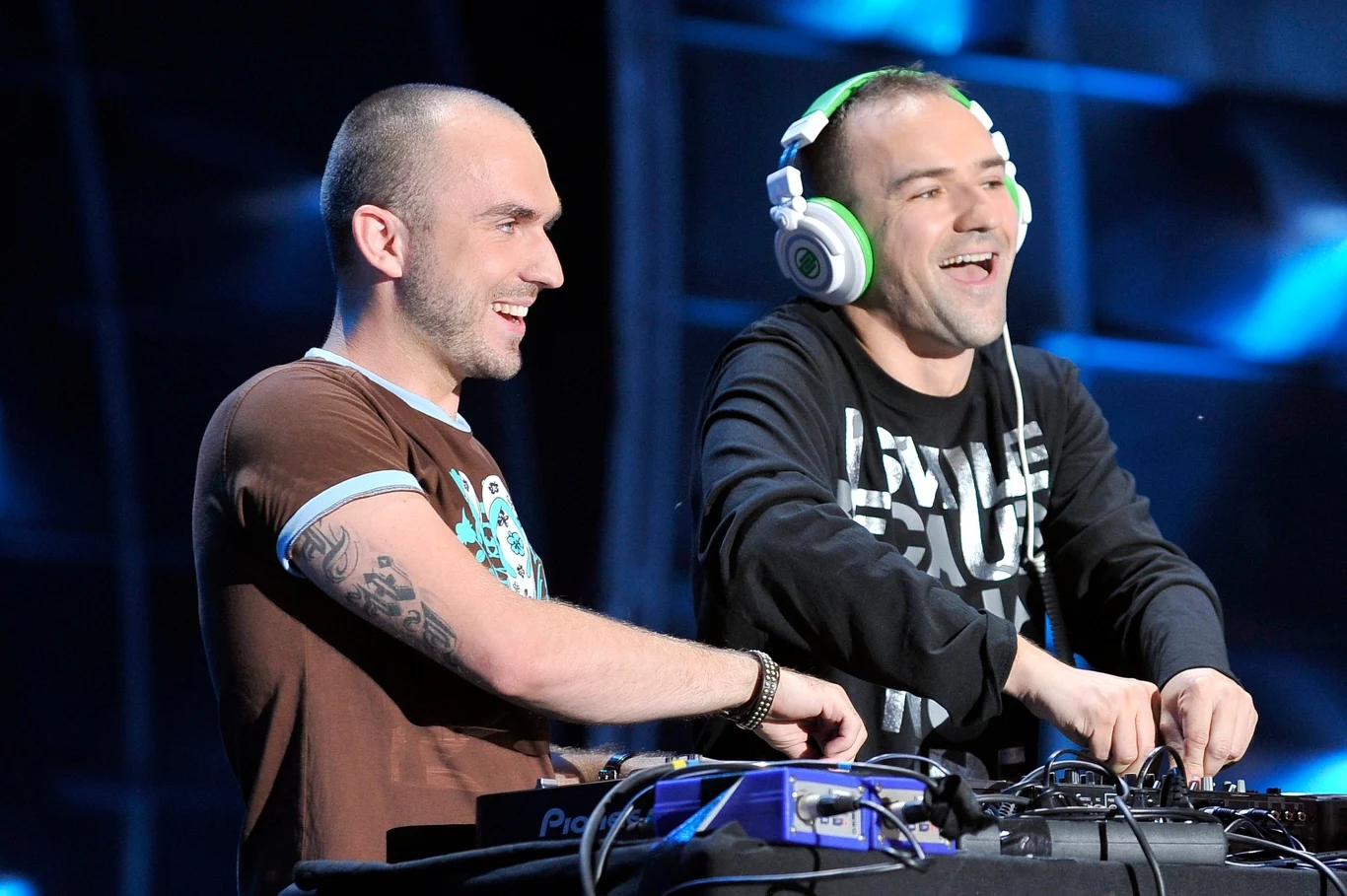 Duet Kalwi & Remi zagra na Roztańczonym PGE Narodowym Dwóch uśmiechniętych mężczyzn stoi przy konsolecie DJ-skiej, jeden z nich ma na uszach słuchawki, obaj wydają się dobrze bawić, wokół niebieskie światła i sceniczne tło.