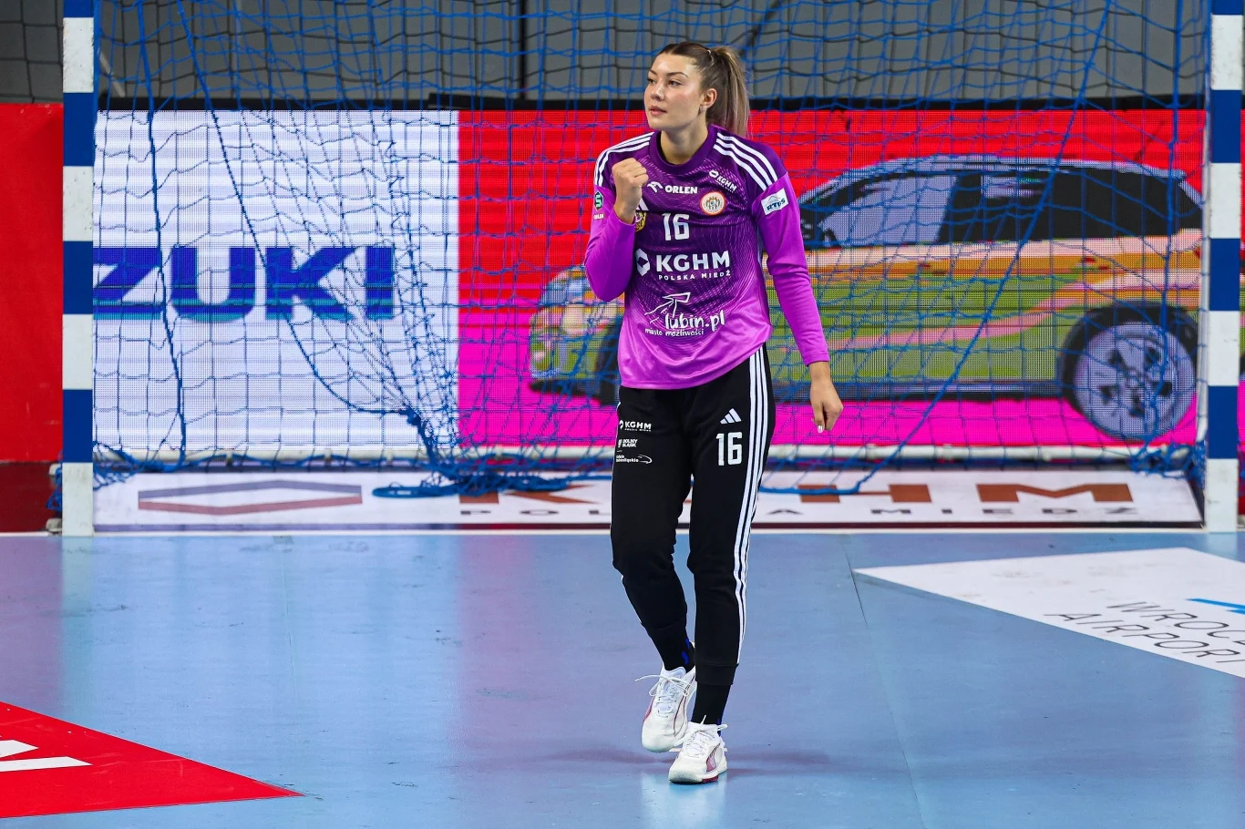 Zawodniczka piłki ręcznej w sportowym stroju bramkarki energicznie świętuje moment podczas meczu, tło stanowi bramka oraz reklama samochodu.