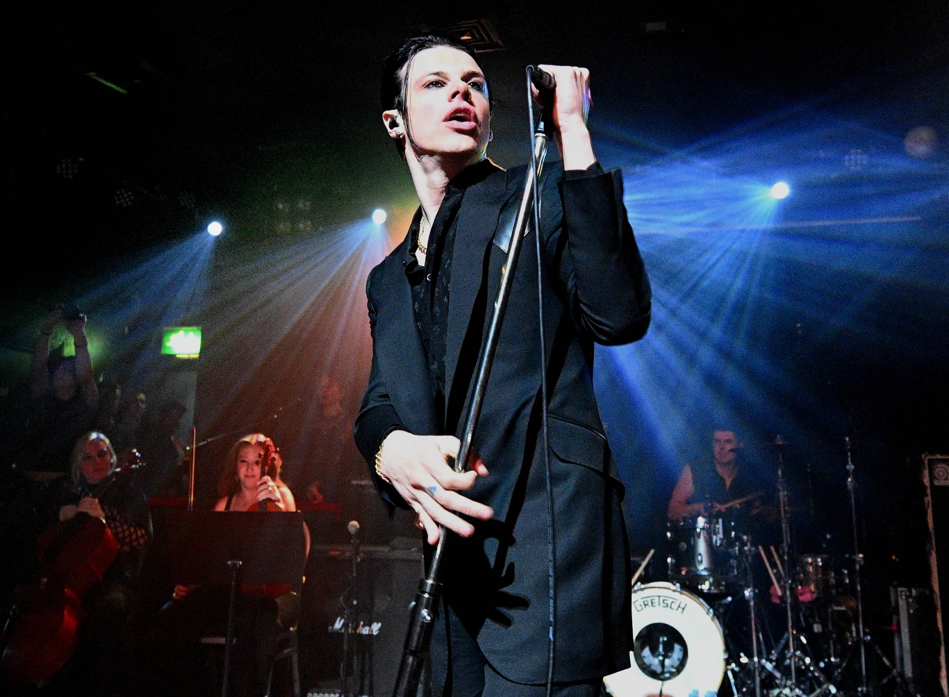 Yungblud 18 października po raz kolejny wystąpi w Polsce Wokalista ubrany na czarno, trzyma mikrofon, występuje na scenie z zespołem instrumentalistów oraz perkusistą, scena oświetlona reflektorami, tło wypełniają muzycy i elementy sceniczne.