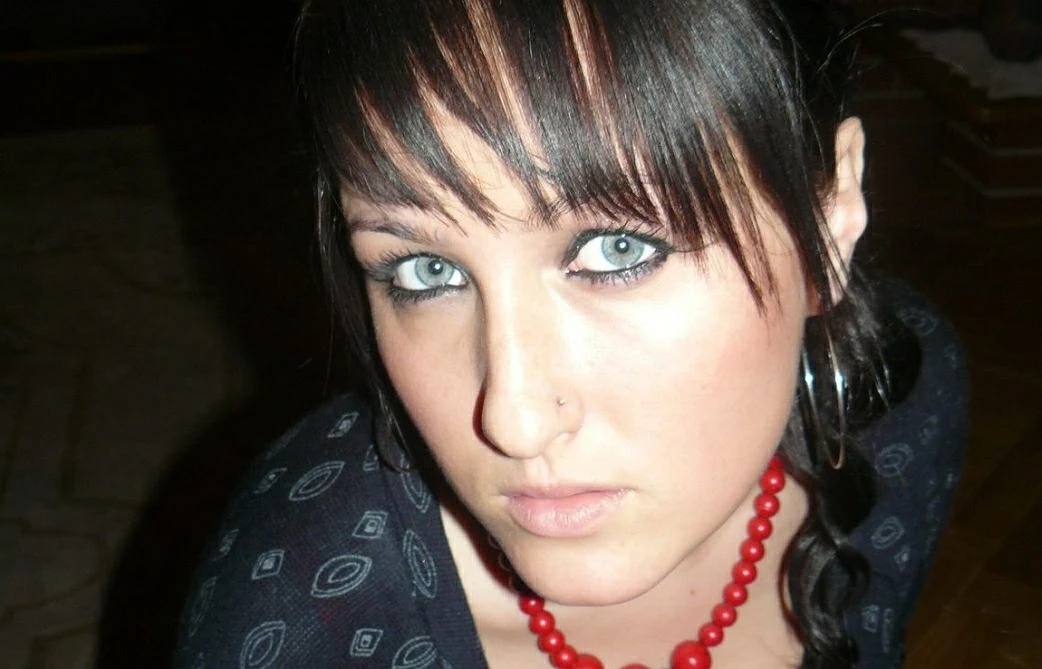 Młoda kobieta z jasnymi oczami, ciemnymi włosami i grzywką spogląda wprost do obiektywu, nosi duże kolczyki koła, czerwony naszyjnik z korali oraz czarną bluzkę z wzorami.