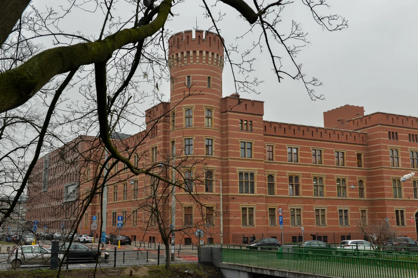 Ceglany, monumentalny budynek o charakterze zamkowy z wieżyczką i wysokimi murami, położony przy ulicy, zimowe drzewa na pierwszym planie, ulica z samochodami i ludźmi.