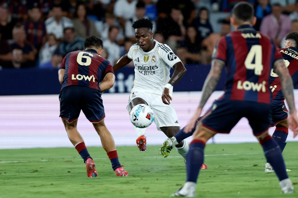 Piłkarz w białym stroju drybluje piłkę, otoczony przez trzech zawodników w granatowo-czerwonych strojach na murawie stadionu podczas meczu piłki nożnej.