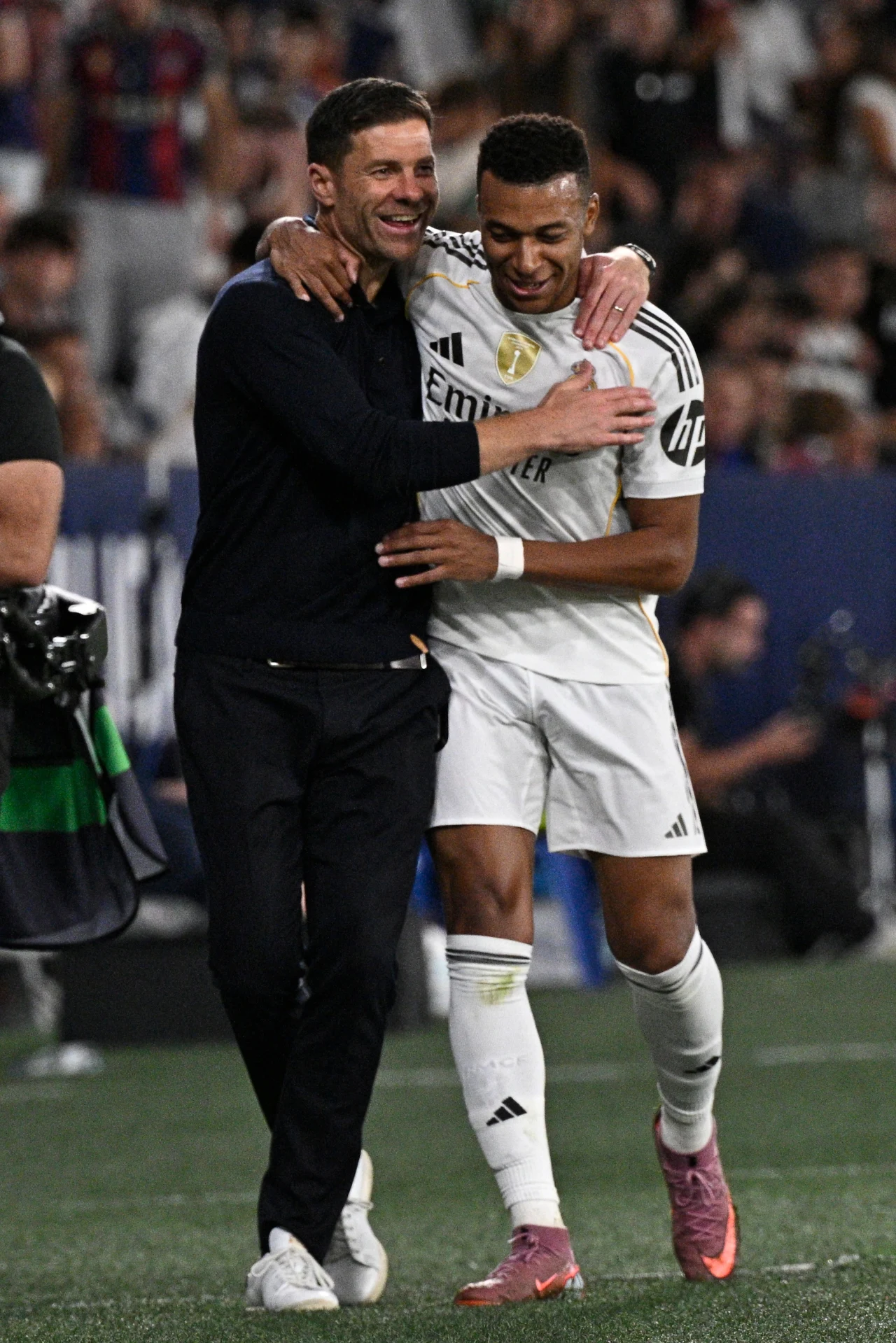 Dwaj mężczyźni obejmują się z uśmiechem na boisku piłkarskim, jeden z nich ubrany jest w strój sportowy drużyny piłkarskiej Realu Madryt, drugi w ciemne ubranie, na trybunach widoczny tłum kibiców.