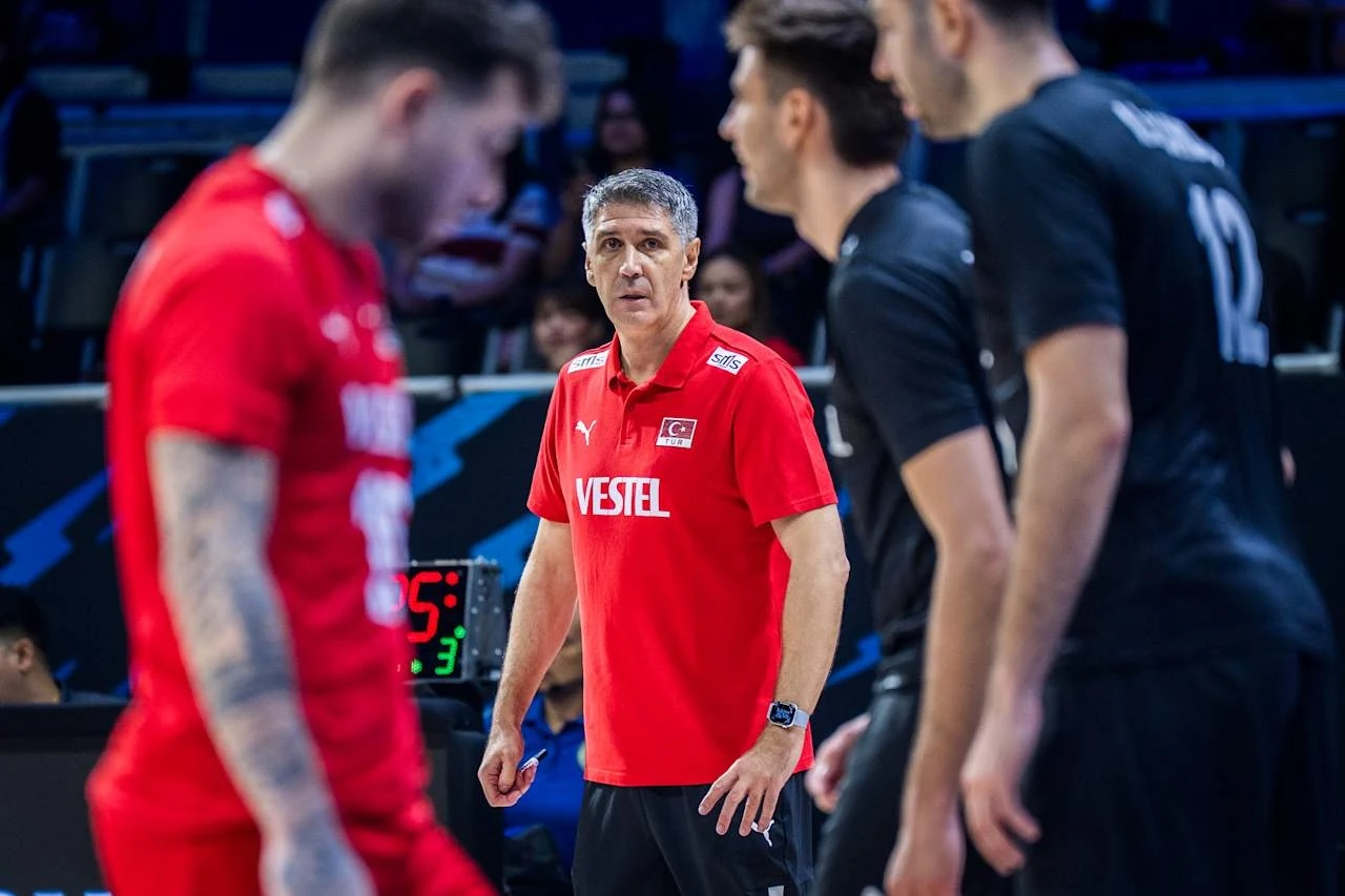 W tle trener reprezentacji Turcji Slobodan Kovac Trener w czerwonej koszulce drużyny narodowej Turcji obserwuje zawodników w trakcie meczu siatkówki, wokół niego znajdują się siatkarze w czarnych i czerwonych strojach, skupieni na grze.