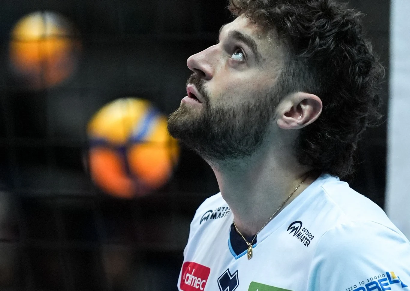 Zbliżenie sportowca w białej koszulce patrzącego w górę podczas meczu siatkówki, w tle niewyraźna piłka siatkowa