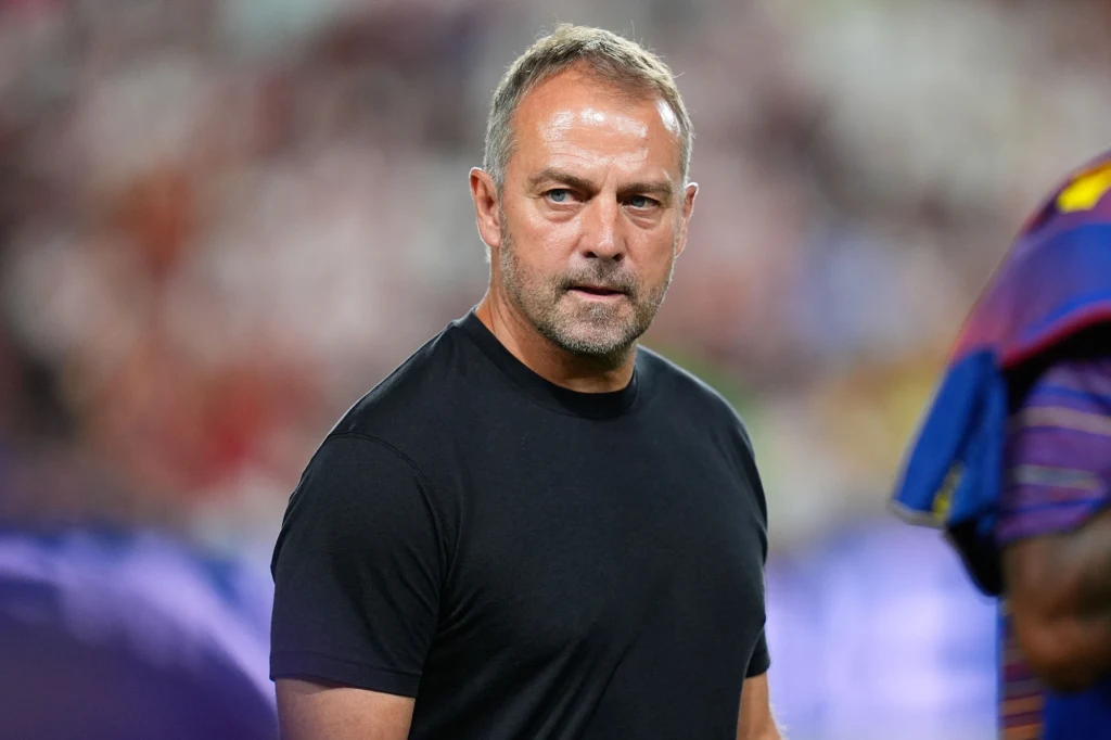 Hansi Flick Mężczyzna w średnim wieku, ubrany w czarną koszulkę, stoi na tle rozmytego stadionu sportowego, ma poważny wyraz twarzy i krótko przycięte włosy, w tle widoczny jest fragment postaci w kolorowej koszulce.