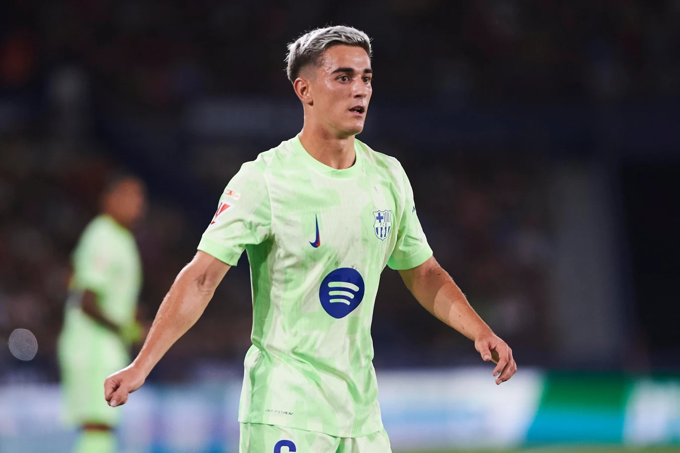 Młody piłkarz w jasnozielonym stroju sportowym z logo sponsorów na stadionie podczas meczu, skupiony na grze, z włosami w odcieniu szarości.