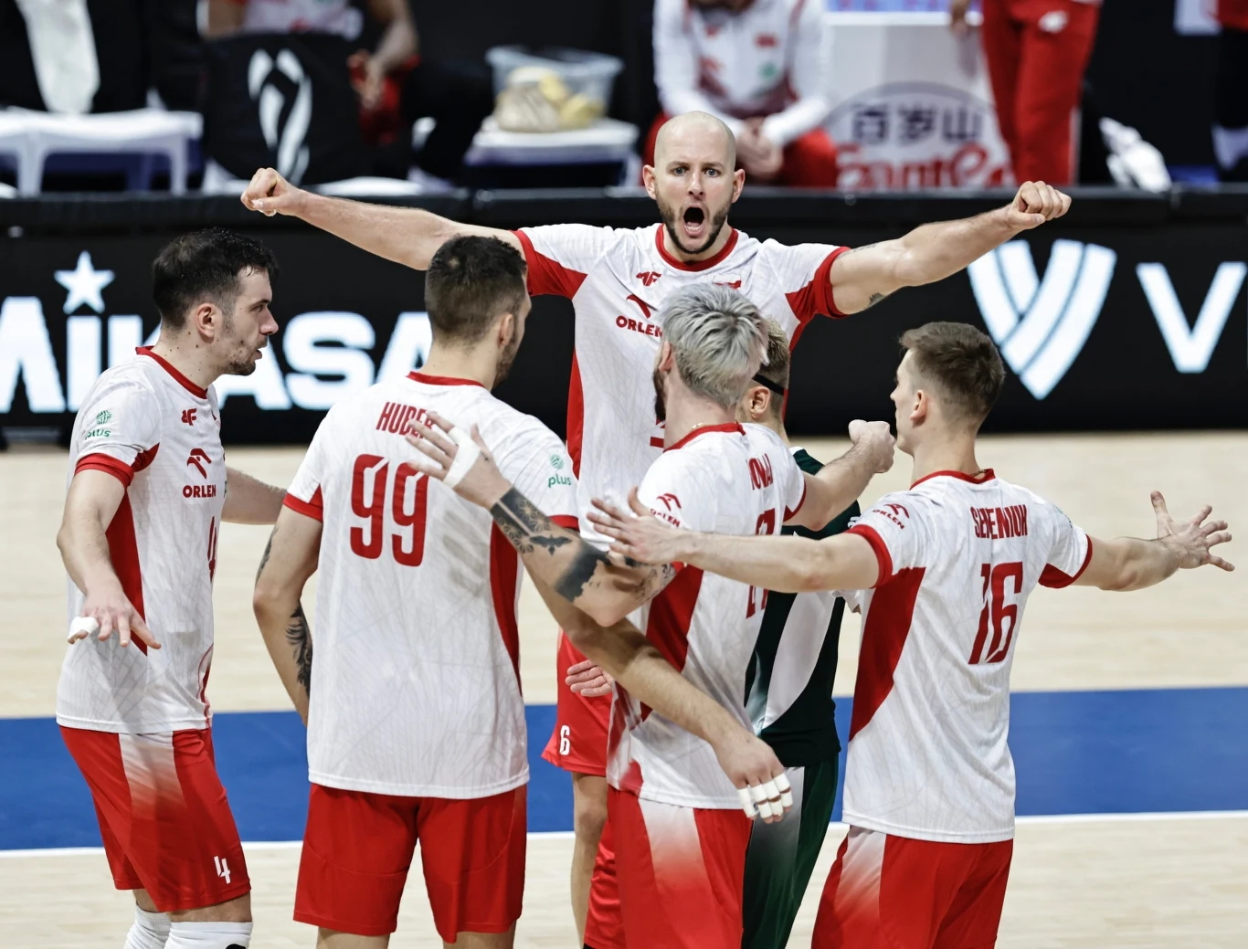 Grupa siatkarzy w biało-czerwonych strojach świętuje zdobyty punkt na boisku, wyrażając emocje poprzez gesty i okrzyki, w tle widoczne elementy hali sportowej.