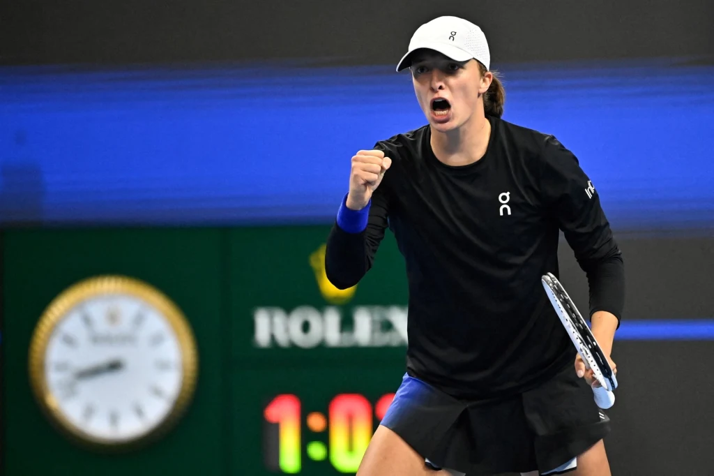 Tenisistka ubrana w sportowy strój wykonuje dynamiczny gest zaciśniętą pięścią, wyrażając emocje podczas meczu na korcie. W tle widoczny zegar i fragment tablicy wyników.